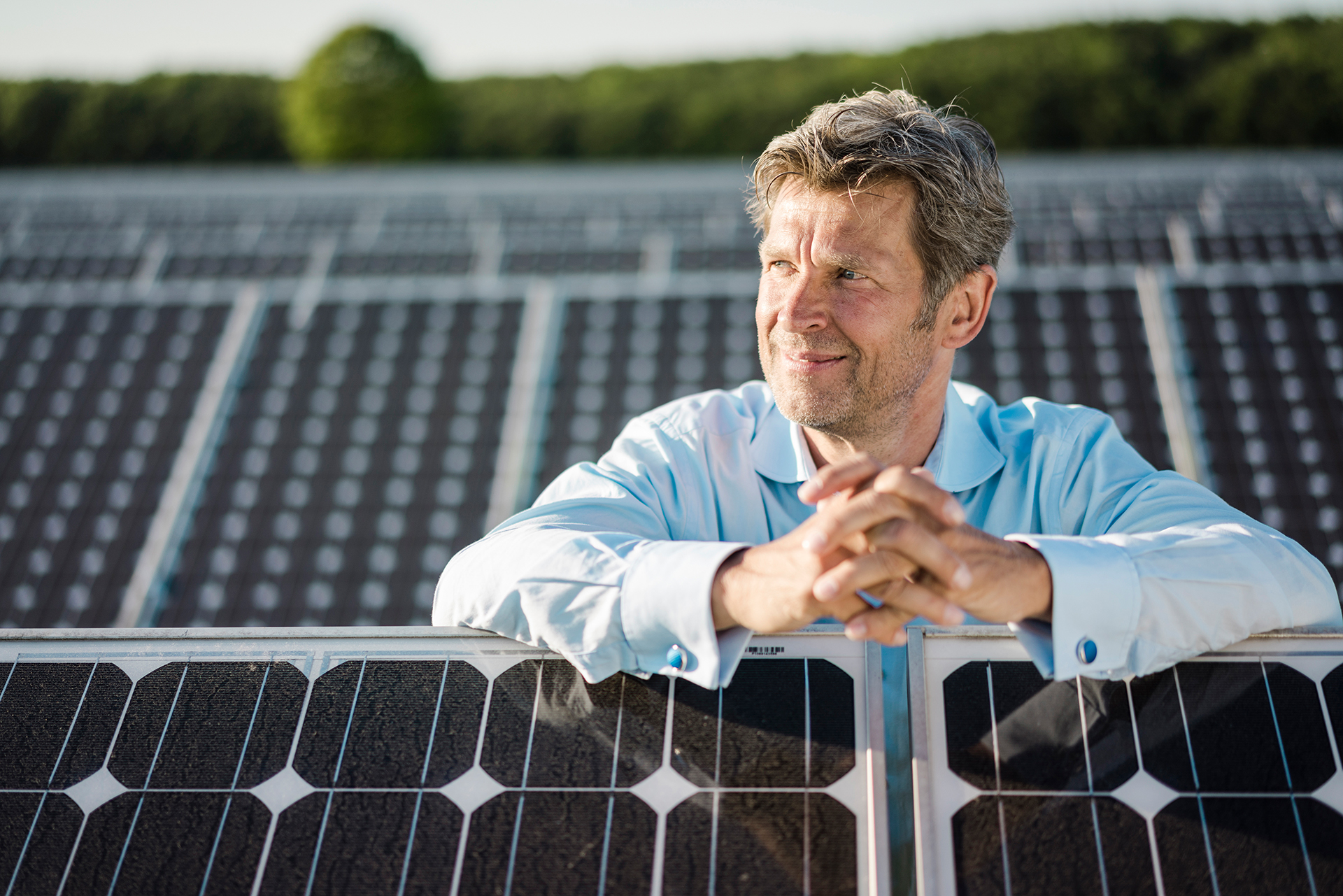 Ein Mann steht zwischen Solarmodulen einer Photovoltaik-Freiflächenanlage. Er hat die Hände verschränkt auf den Modulen abgelegt und schaut seitlich in die Sonne.