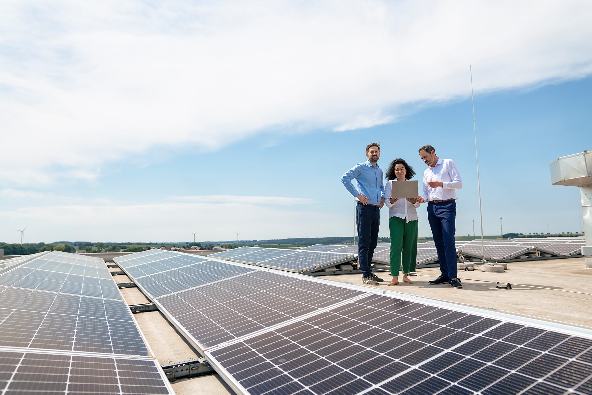 Drei Personen, zwei Männer und eine Frau, stehen auf einem Dach. In der linken Bildhälfte sind Solarmodule auf dem Flachdach leicht schräg aufgestellt.