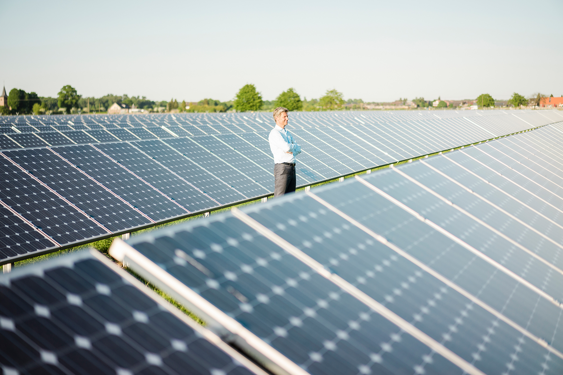 Ein Mann steht zwischen Solarmodulen einer Photovoltaik-Freiflächenanlage. Im Hintergrund vereinzelt Bäume und Gebäude.