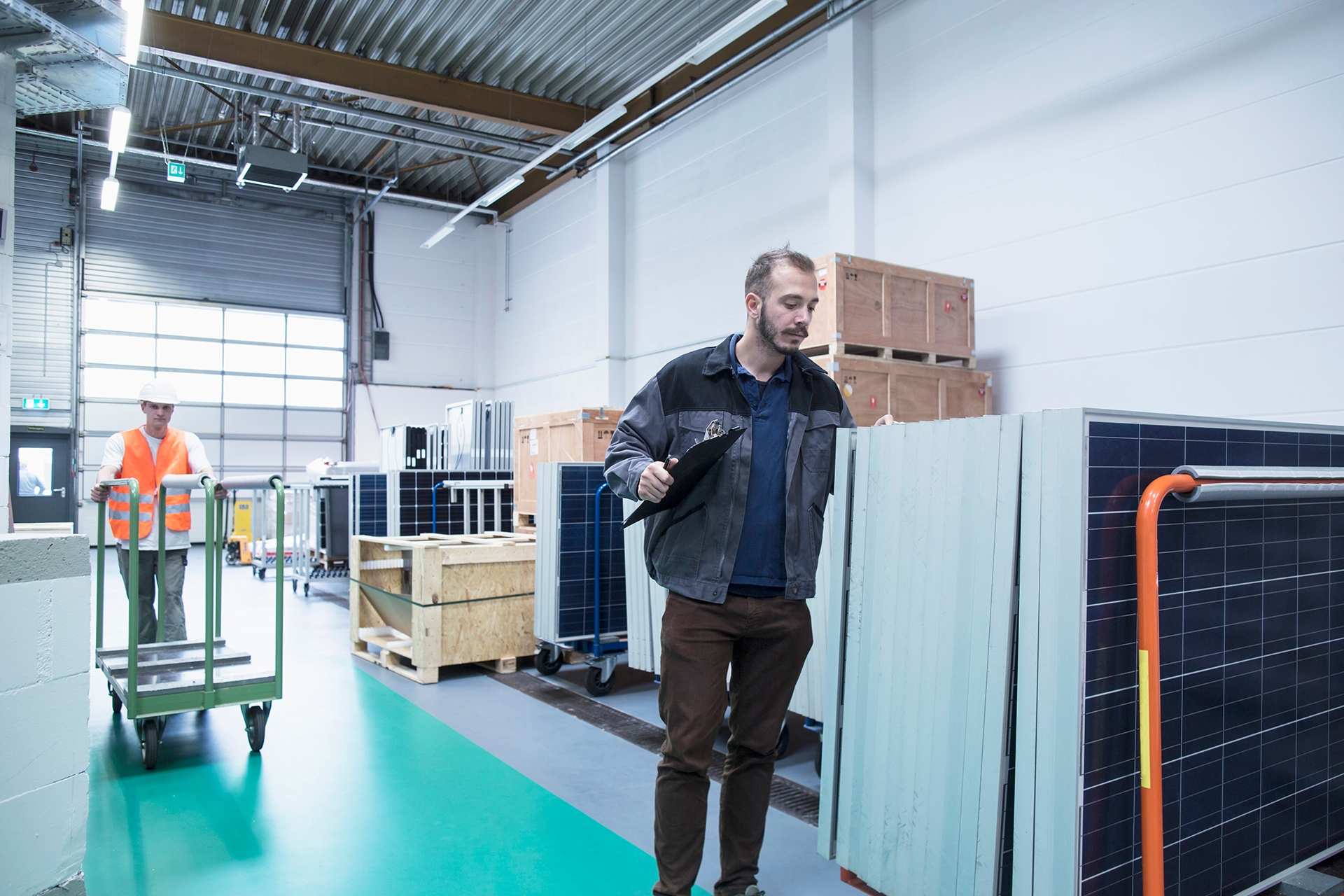 Zwei Männer stehen in einem Lager. Einer schiebt einen Rollwagen, ein anderer kontrolliert mit einem Klemmbrett Solarmodule.