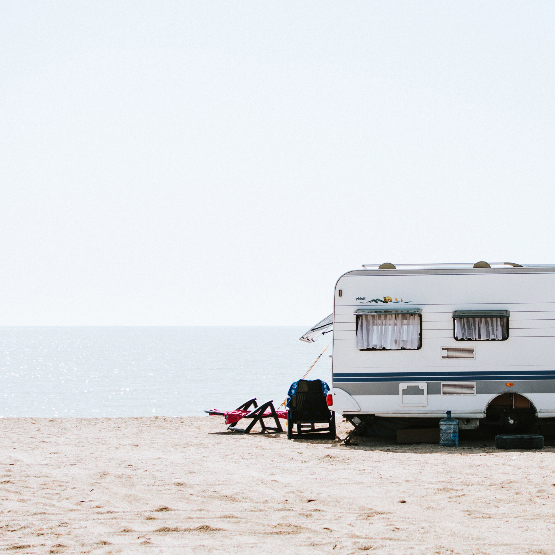 Ein Wohnwagen steht an einem Strand. Neben dem Wohnwagen sind zwei Liegestühle aufgestellt.