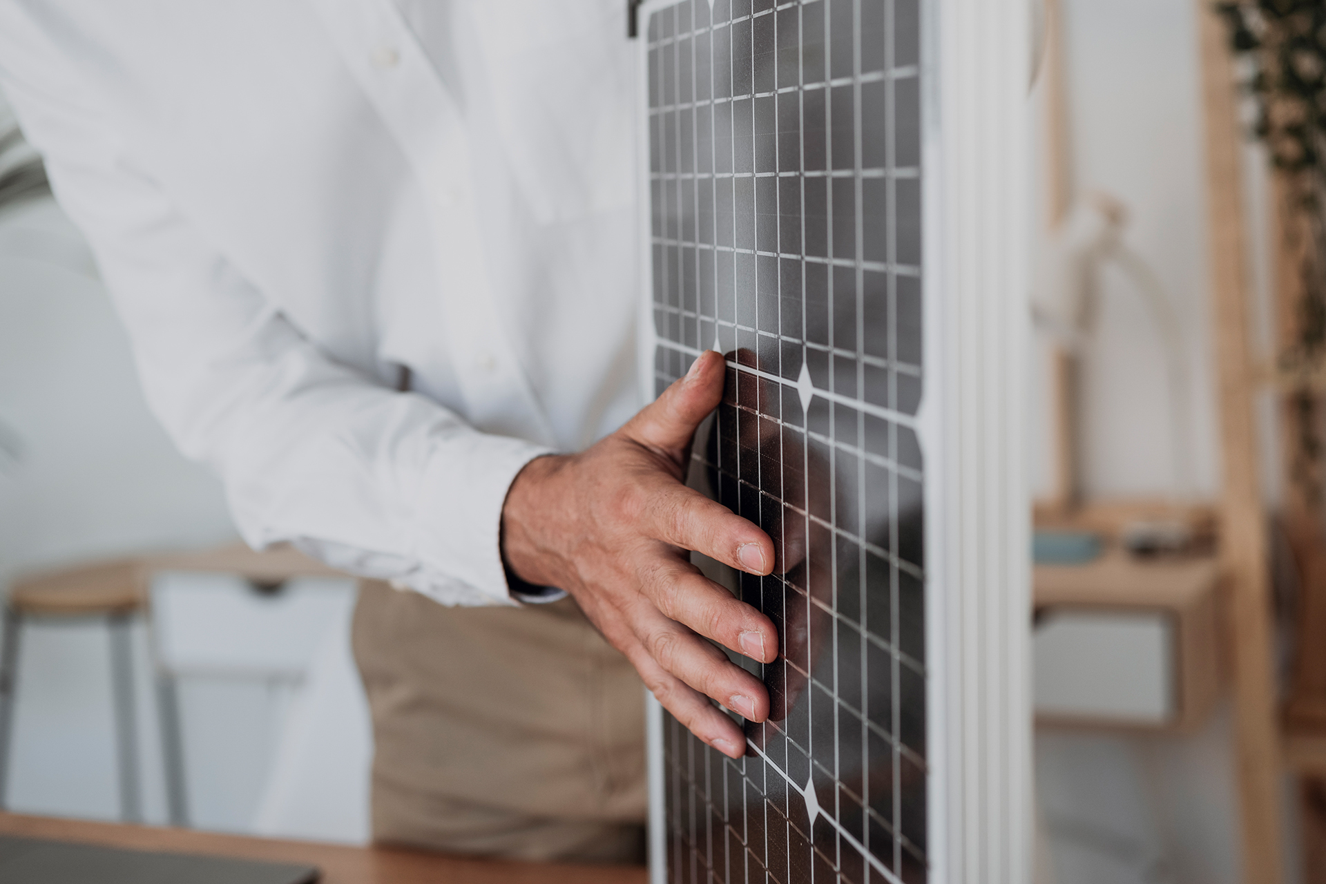 Eine Person mit weißem Hemd und brauner Hose streicht mit der Hand über ein Solarmodul. Im Hintergrund sieht man Holzmöbel vor weißen Wänden.