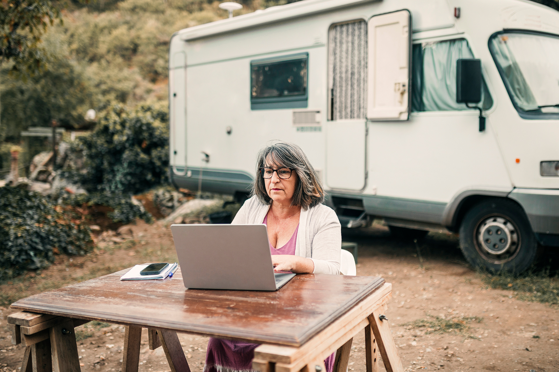 Im Hintergrund steht ein Camper-Van in der Natur. Im Vordergrund sitzt eine Frau mit grau-schwarzen Haaren an einem Holztisch vor ihrem Computer.